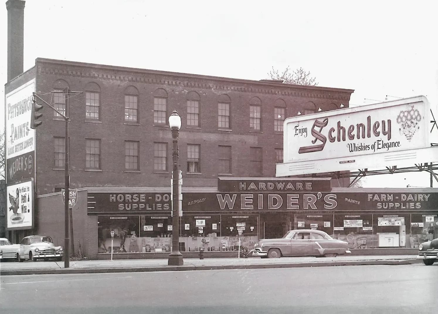 Black-and-white photo of Historic black-and-white photo of the original Weiders Hardware store buildinga three-story brick commercial building on a street corner. A long storefront awning reads "WEIDER'S HARDWARE," flanked by "HORSE-DOG SUPPLIES" on the left and "FARM-DAIRY SUPPLIES" on the right. A billboard on the roof advertises Schenley whiskey. Four 1950s cars are parked along the curb.